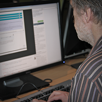 En mann med skjegg og stripet skjorte sett bakfra - han sitter konsentrert ved en PC-skjerm