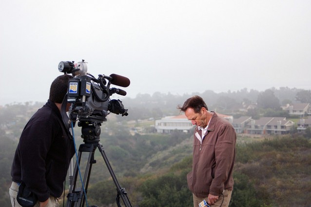 En reporter stående foran et videokamera med bøyd nakke. I bakgrunnen grålys, landskap, noen bolighus. Jordskjelv i California
