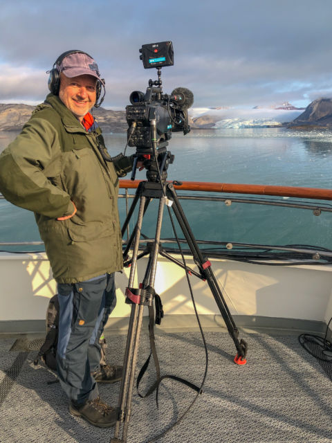 En fotograf står sammen med et kamera på dekket av et skip. En isbre i bakgrunnen. 