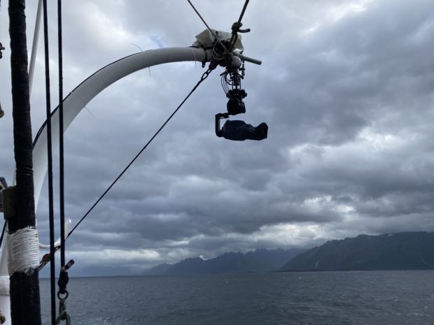 Et kamera henger fra en kran-lignende Davit som vanligvis brukes til å feste livbåter og annet til en båt. Skyer, høye fjell og hav i bakgrunnen.