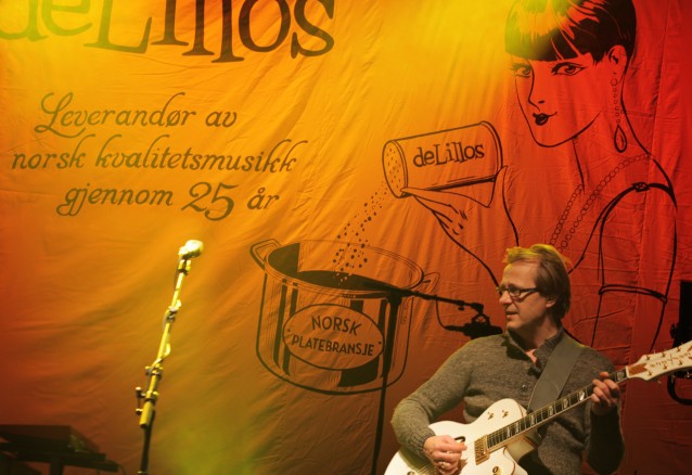 Lars Lillo-Stenberg står på scenen i hverdagsklær og spiller gitar. Bak ham en backdrop med teksten deLillos Leverandør av kvalitetsmusikk i 25 år. Ved siden av en stilisert kvinne som drysser fra en boks merket deLillos ned i en gryte merket norsk platebransje