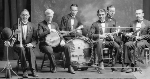 Six musicians pose in front of a backdrop with their instruments, including saxophones, clarinets, a banjo, drums and trumpet with mutes. The drum has been painted with a seascape and the word "Orchestra" in faux Oriental letters.  Ca 1925.