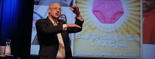 Clay Shirky står på scenen og snakker foran et bilde som blant annet viser en stor rosa dametruse