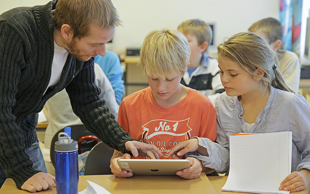 Skolesituasjon. En mannlig lærer lener seg over to elever og viser dem noe på iPaden de sitter med.