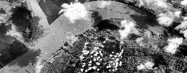 Luftfoto av en by ved en elv. Røykdotter stiger opp fra bombede områder