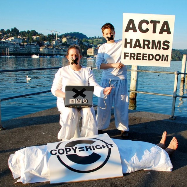 To hvitkledte aktivister med et skilt med teksten "ACTA harms freedom", sorte kryss av tape over leppene, sort kryss på en laptop og et "lik" under et hvitt laken med teksten "Copy = Right"