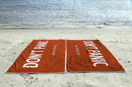 To håndklær liggende på en strand, begge påtrykt teksten DON'T PANIC