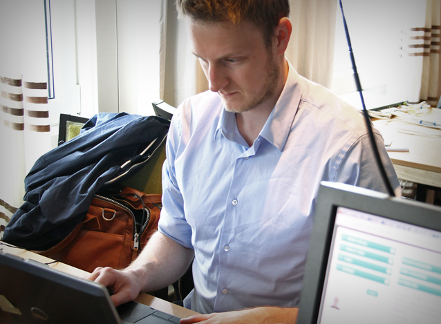 Sett over hjørnet av en PC: En mannlig journalist i blå skjorte sitter konsentrert over en bærbar PC. ved siden av ham en sekk og en jakke.