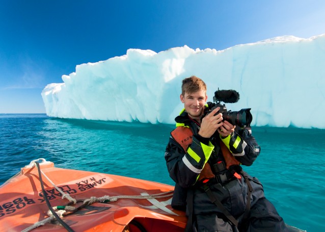 En person i lettbåt ved et isfjell, holdende på et speilreflekskamera