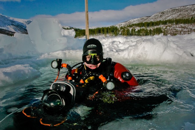 En dykker med et kamera i undervannshus stikker hodet opp fra et hull i isen