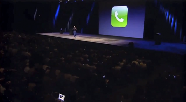 Skjermskudd fra keynote-video, kamera langt bak i salen. Steve Jobs er fyrstikk-stor på scenen, projeksjonen bak ham viser iPhonens telefonikon, i salen fullt av publikum og lyset fra to-tre laptoper