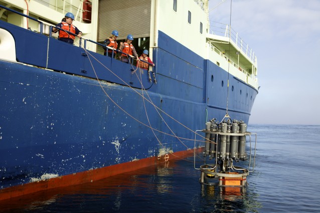 et bilde som viser et blått forskningsskip hvor forskere står langs ripen og lårer et instrument ned i havet.