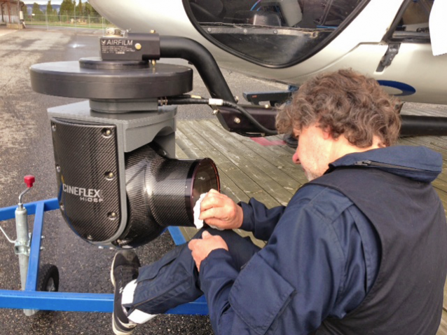 En mann pusser av frontglasset på et kamera montert på en arm på siden av et helikopter