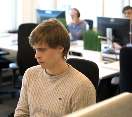 Kristoffer Lium sitter ved pulten sin i det åpne landskapet i NRK. To kollegaer sitter og jobber i bakgrunnen.