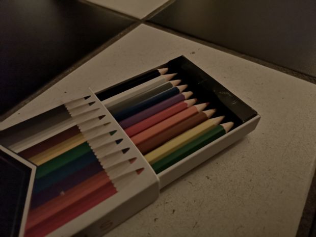 Bilde av en boks fargeblyanter. Svake farger og tydelig tatt i lite lys.