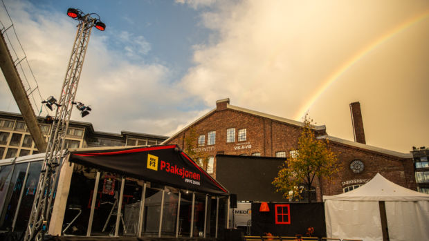 Teltet til P3aksjonen på Solsiden i Trondheim foran en murbygning med en regnbue på skyet himmel, med lysrigg i forgrunnen