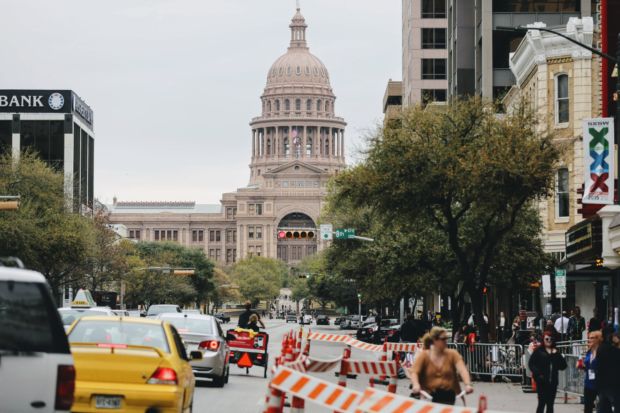 En gate med trafikk og veisperringer i forgrunnen, med Texas State Capitol i bakgrunnen. En bygning med et høyt tårn i beige farge, som ligner mye på den amerikanske kongressen.