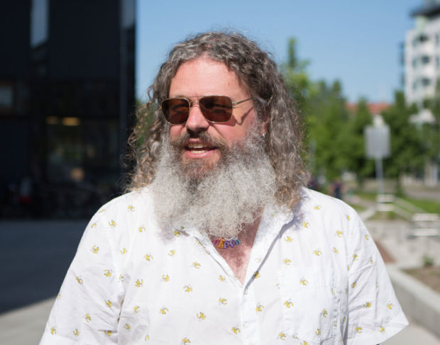 En mann med langt hår, langt skjegg og solbriller står midt i en gate.
