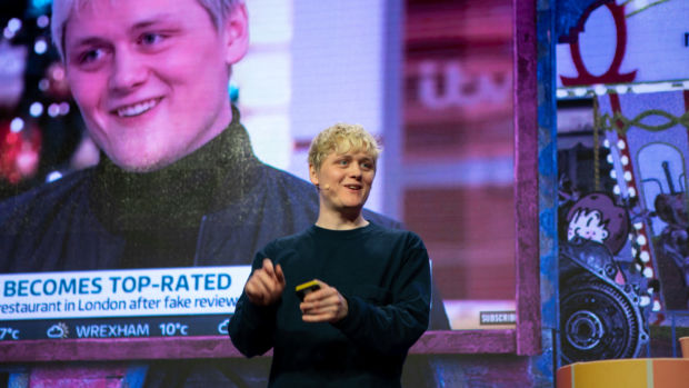 En ung mann med bleket hår står på en scene med en presentasjons-fjernkontroll i hånden. Han har på svart genser. Bak han vises det fram et enormt skjermbilde fra en slags nyhetssending. Det er av han selv sittende i et TV-studio hos britiske ITV, med teksten "Ble topplistet" synlig.