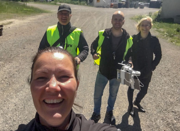 En kvinne tar en selfie med tre personer i bakgrunnen: to i gule vester som markerer at de jobber med dronefoto. 