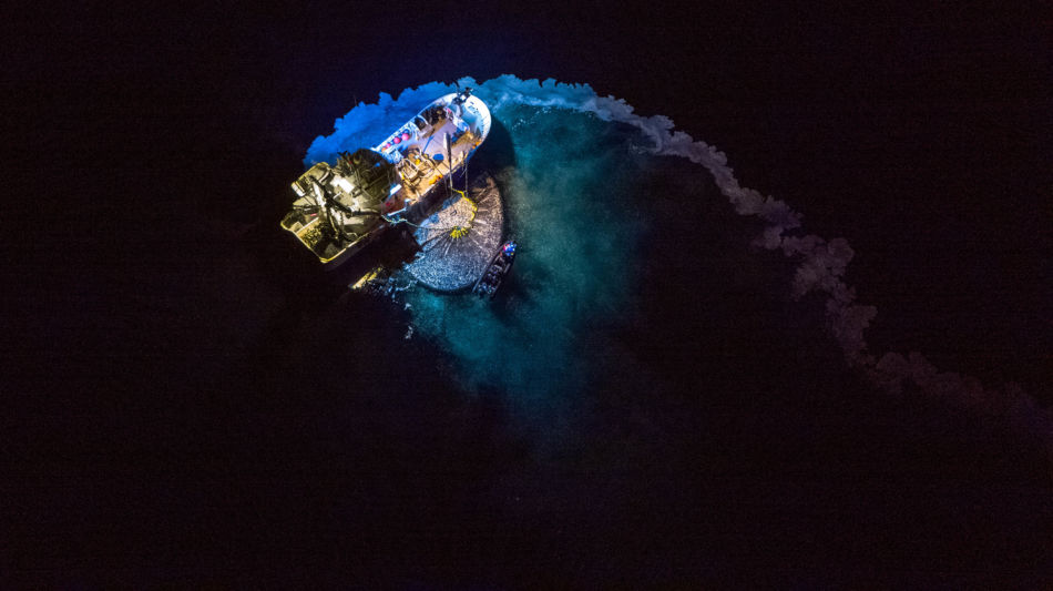 En fiskebåt med trål sees ovenfra. Den ligger på et flatt hav, midt på natten. Trålen er snurpet sammen, og det ligger en hale av fiskeolje etter båten. Sjøen er delvis opplyst av båtens lamper, og ser grønn, lilla og blå ut. 
