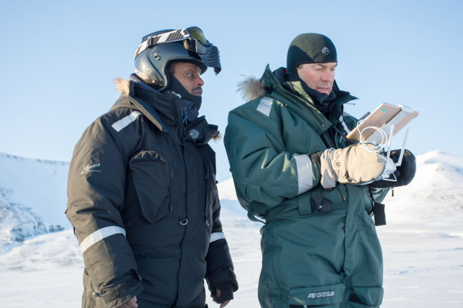 To personer i scooterdresser står i et snødekt landskap på Svalbard. Person 1 følger med på person 2 som har en fjernkontroll i hendene og flyr drone. 