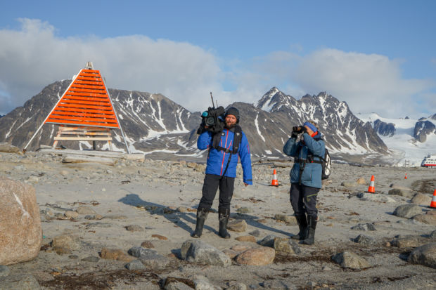 En fotograf med TV-kamera på skulderen og programleder Kari Toft med kikkert står på en øde, steinete strand på Svalbard