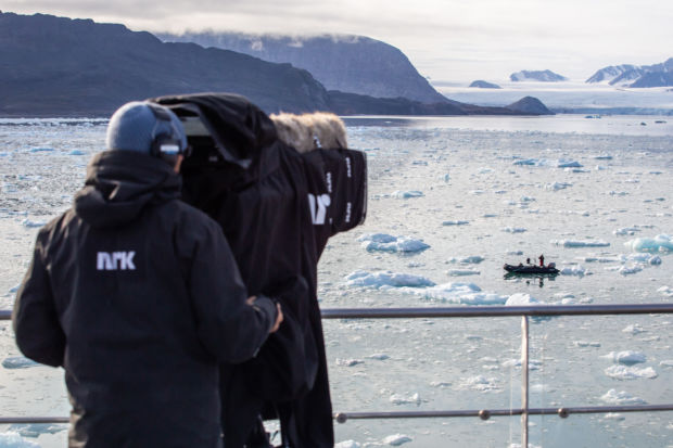 En person på dekket av en båt med telekamera. En liten båt på sjøen i bakgrunnen, med små isklumper i sjøen og en isbre i bakgrunnen. 