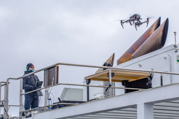 En mann står på et båtdekk like ved en skorstein og lander en drone på en plattform. 