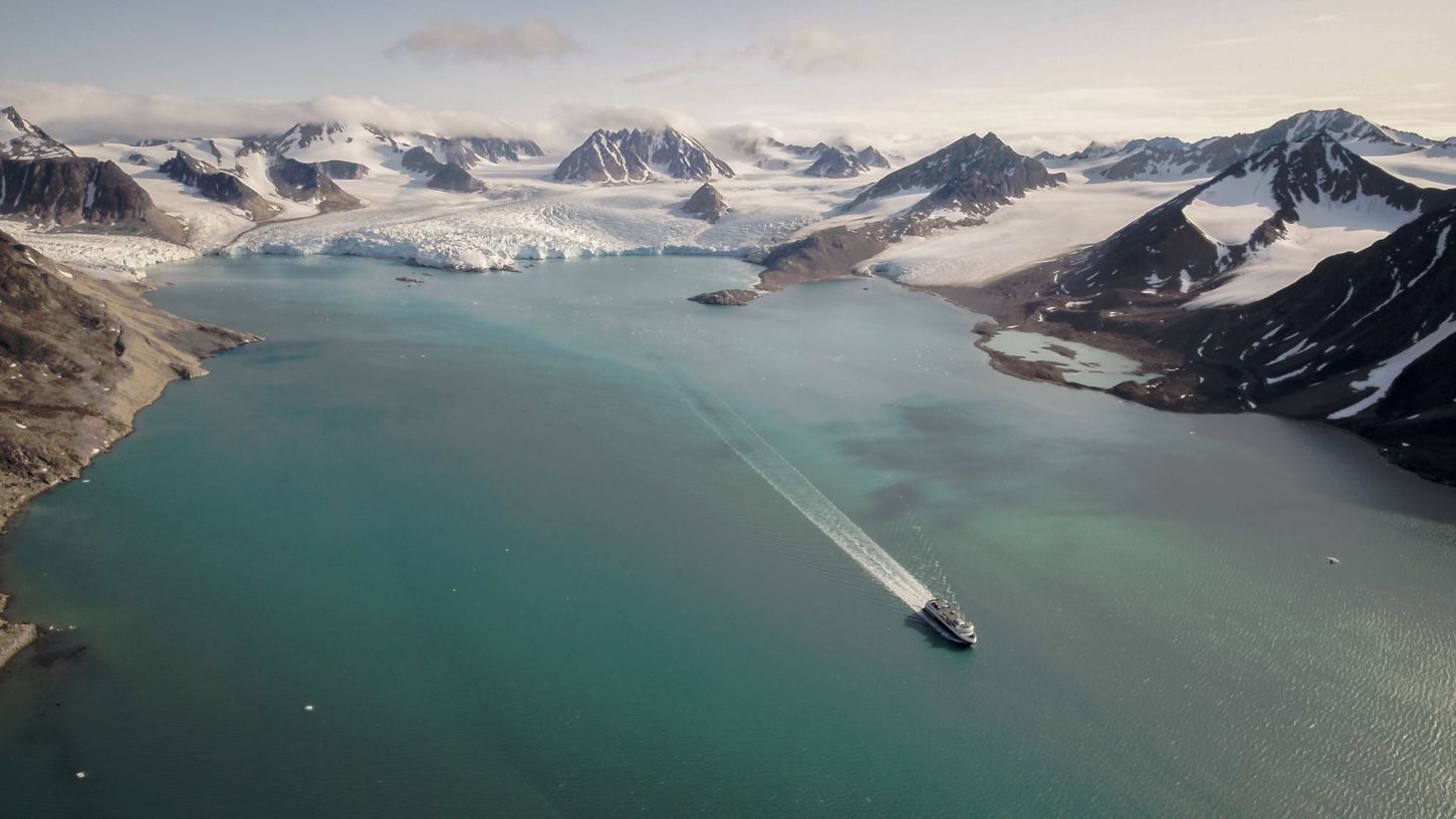 Et stort og vidt bilde av en isbre med en fjord foran. I ene bildekant et skip som seiler bort fra breen.