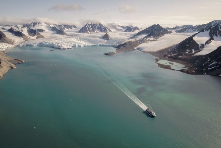 Et stort og vidt bilde av en isbre med en fjord foran. I ene bildekant et skip som seiler bort fra breen.