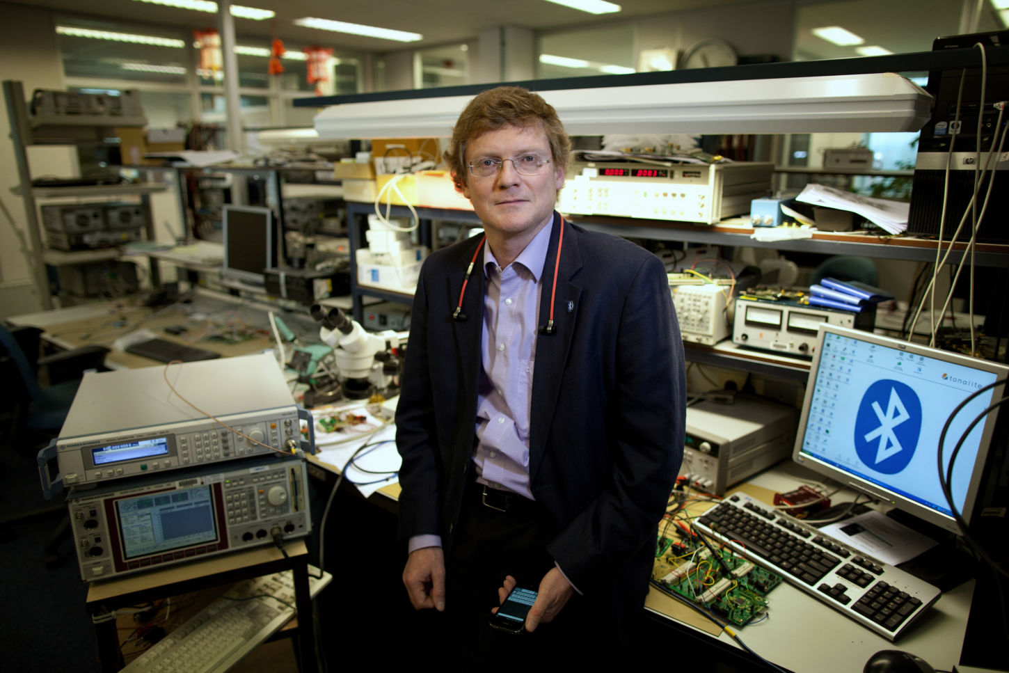 En mann sitter på et arbeidsbord i et teknisk laboratorium. Det er flere apparater og datamaskiner i bakgrunnen, og på en av skjermene vises Bluetooth-logoen.