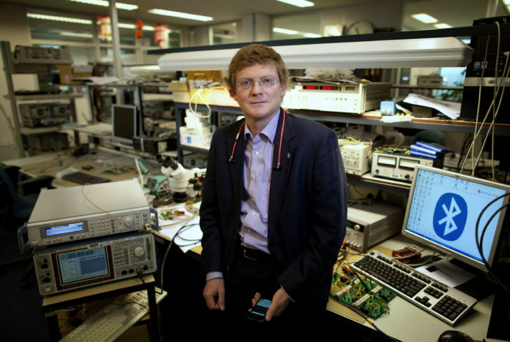 En mann sitter på et arbeidsbord i et teknisk laboratorium. Det er flere apparater og datamaskiner i bakgrunnen, og på en av skjermene vises Bluetooth-logoen.