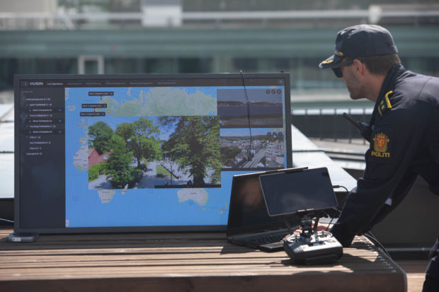 En politibetjent står vendt mot en stor skjerm som viser et kart med flere bilder over. Bildene er fra store norske byer som Trondheim og Tromsø.
