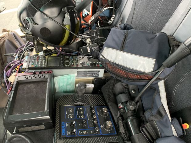 Et nærbilde av et stolsete med kamerakontrollere og skjermer. En hjelm og et munnbind ligger også synlig. Det er litt kaotisk med utstyr og kabler liggende rundt.
