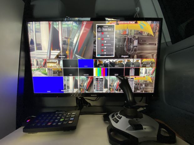 En skjerm med mange ulike kamerakilder på er i fokus. Like under, på en treplate står en sort lav boks og en joystick.
