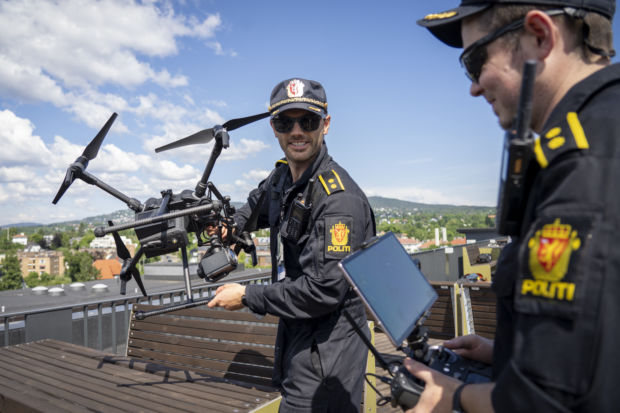 To personer i politiuniform er avbildet på et tak i Oslo. Den ene holder en stor drone og ser inn i kameraet. Den andre holder en styringsenhet med et påmontert nettbrett og ser ned på denne skjermen.