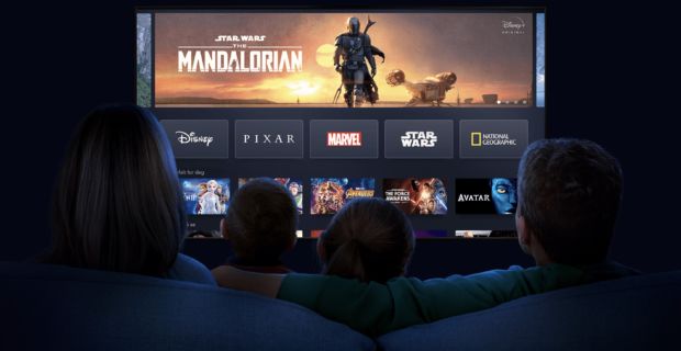 En stor TV-skjerm med Disney-serier som The Mandalorian øverst og logoer som Marvel og National Geographic. I forgrunnen kan man se silhuettene av en mor, far og to barn som sitter i en sofa. Disse er sett bakfra og TVen er i fokus.