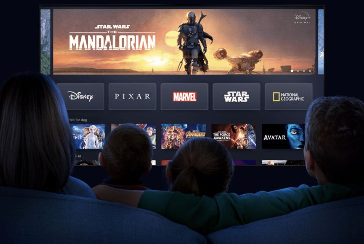 En stor TV-skjerm med Disney-serier som The Mandalorian øverst og logoer som Marvel og National Geographic. I forgrunnen kan man se silhuettene av en mor, far og to barn som sitter i en sofa. Disse er sett bakfra og TVen er i fokus.