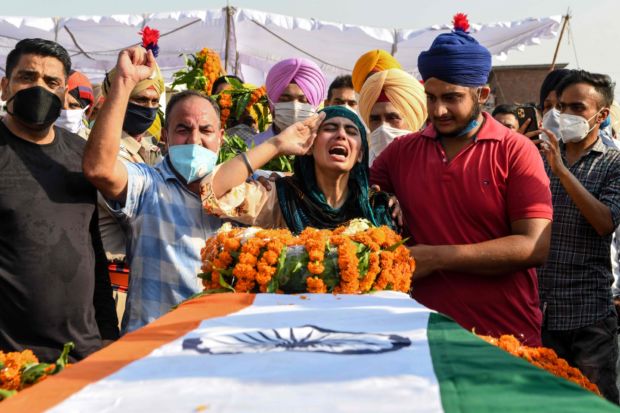 Et begravelsesfølge ikledt munnbind står rundt en kiste drapert i det indiske flagget med blomster på.