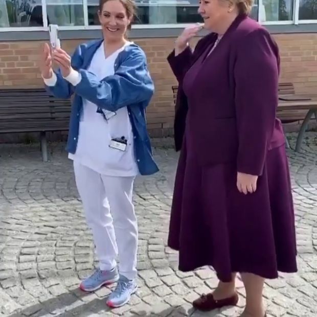 En person i sykehus-klær står ved siden av norges statsminister som har på en lilla kjole. Førstnevnte holder opp en mobiltelefon som begge ser på.