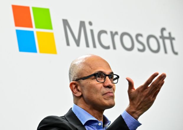 En person med briller og dress gestikulerer med hånden foran en Microsoft-logo i bakgrunnen.