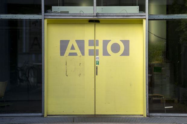 Et nærbilde av en gul skyvedør med bokstavene AHO, som er omkranset av et moderne bygg av glass og stål.