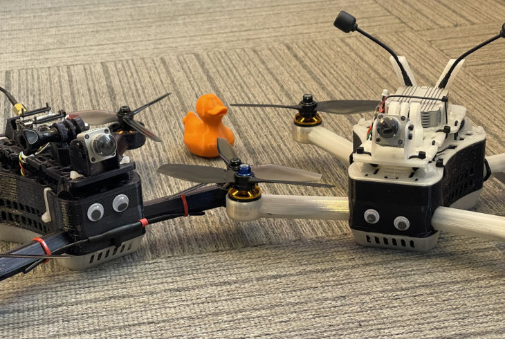 To 3D-printede droner står på gulvet med påklistra øyne med en oransje NRKbeta-and i bakgrunnen