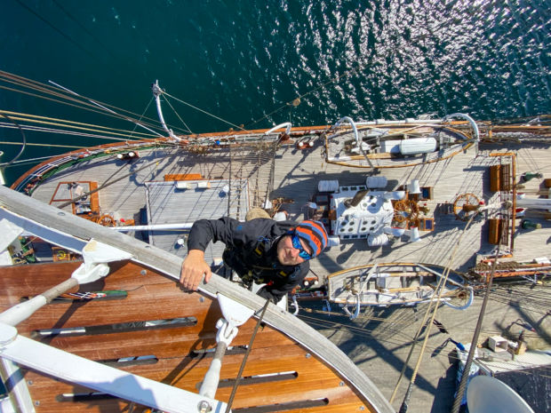 Bilde fra toppen av masten og ned på dekket av en seilskute. En smilende mann er på vei opp masten mot kameraet.