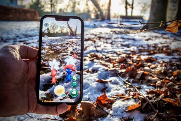 Bilde av mobiltelefon som viser spillet Pikmin Bloom mot snø og høstløv