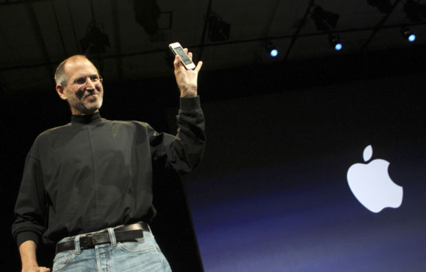 En mann står på en scene med en stor Apple-logo i bakgrunnen og holder opp en mobiltelefon. Han er ikledd en sort høyhalset genser og dongeribukser med et sort belte.