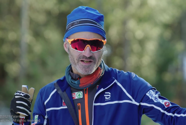 A man in full blue ski gear, with logos of Norwegian companies visible.